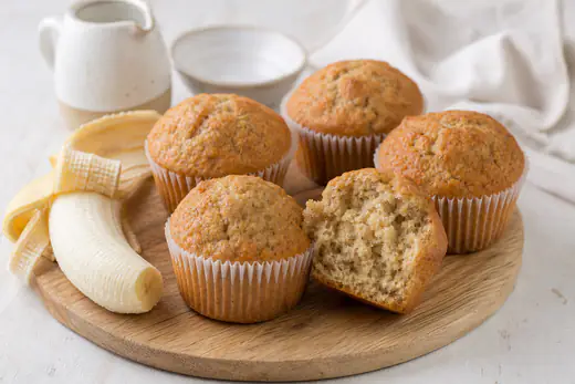 Muffin alla banana con la cupola dorata nei pirottini, accanto a una banana matura sbucciata e a un muffin tagliato a metà