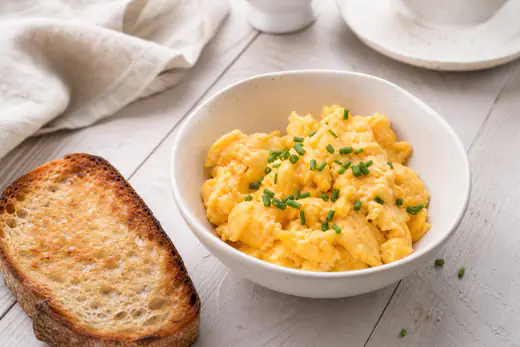 Uova strapazzate soffici e giallo chiaro in una ciotolina bianca, con erba cipollina e una fetta di pane tostato accanto